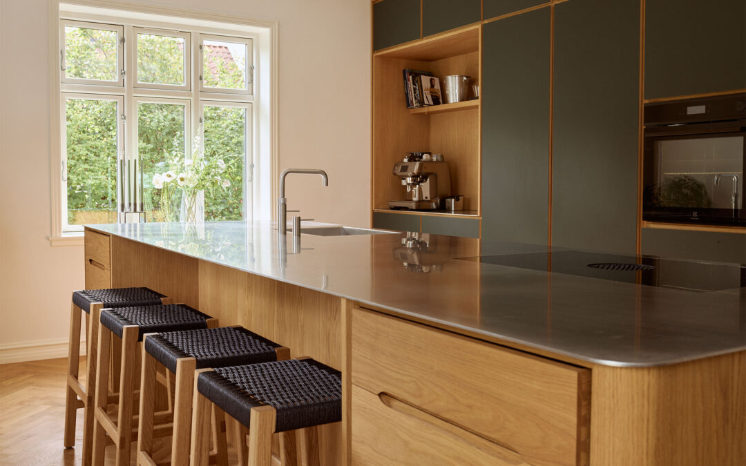 Moderne snedkerkøkken med en stor køkkenø i eg, stålbordplade og fire barstole med sort flet, foran olivengrønne skabselementer og et lyst vindue.
