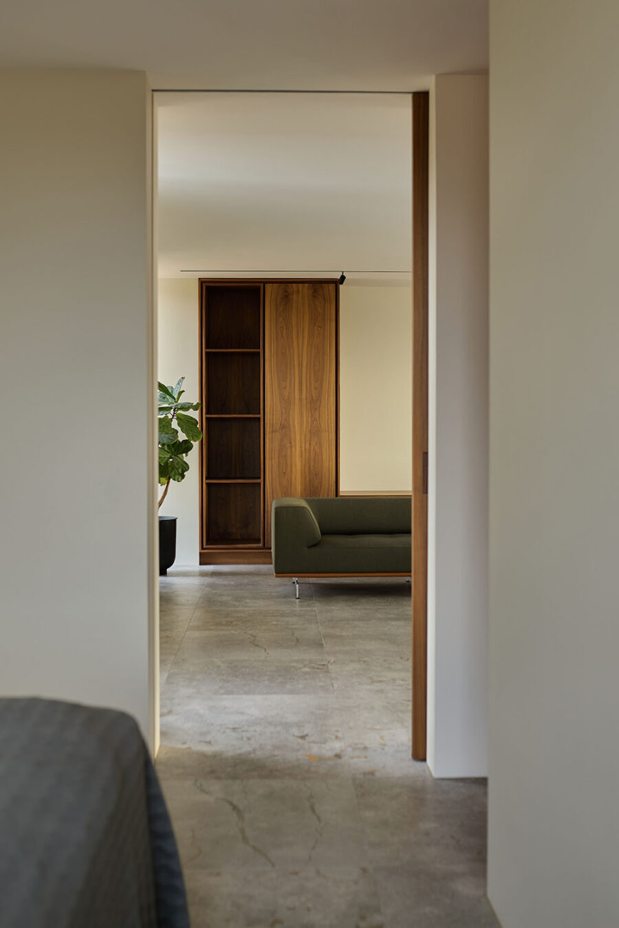 Kig gennem døråbning til minimalistisk stue med snedkerskab i valnød, olivengrøn sofa og naturstensgulv.